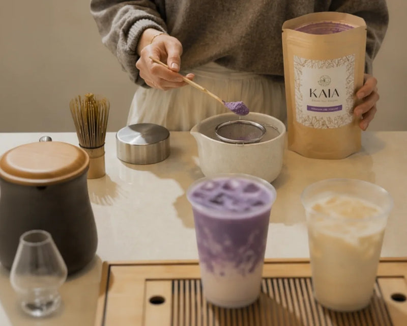 Person preparing a KAIA Origin ube latte with chashaku and sieve, kraft pouch and matcha whisk on a wooden bar